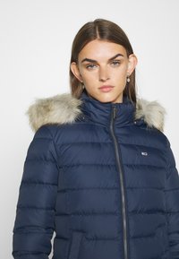 Chaqueta de plumas azul marino con cierre de cremallera y bolsillos laterales. Presenta una capucha con forro de piel sintética y un textura acolchada. Parche del logo en el pecho.