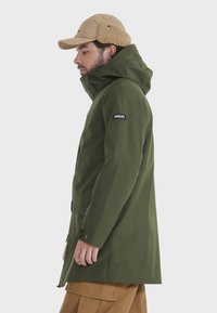 Olivgrüne wasserdichte Jacke mit Kapuze, ausgestattet mit einem schwarzen Aigle-Logo am Ärmel. Das Design umfasst verstellbare Bündchen und einen geraden Schnitt.