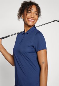 Námornícky polo tričko s klasickým límcem, krátkými rukávy a třemi knoflíky. Hladká látka s pohodlným střihem. Golfová hůl opřená o rameno.