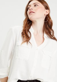 Chemise blanche à manches longues avec un tissu léger et texturé ; dotée de deux poches poitrine et d'un col décontracté.