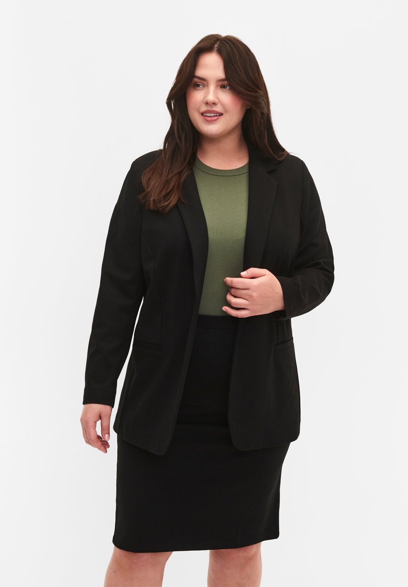 Blazer nero con due tasche frontali, abbinato a un top verde a costine e una gonna a tubino. Texture liscia, vestibilità sartoriale e aspetto classico.
