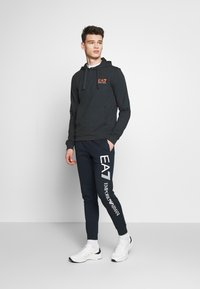 Melns krekls ar oranžu logo un priekšējo kabatu, savienots ar tumši ziliem jogger šortiem ar baltiem uzrakstiem. Modelis valkā baltas sporta apavus.