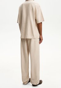 Beżowa lniana koszula z krótkim rękawem i luźne spodnie, obie wykonane z miękkiego materiału. Koszula ma prosty fason i prosty krój.