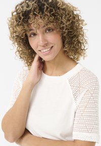 T-shirt blanc à rayures avec des manches courtes en dentelle. Texture douce et col rond. Le mannequin pose la main sur le cou en souriant.
