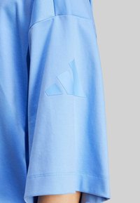 Svetlomodrá košeľa s krátkym rukávom, mäkká na dotyk, s diagonálnym logom troch pruhov v tmavšej modrej farbe na ľavom rukáve. Čistý dizajn, okrúhly výstrih.