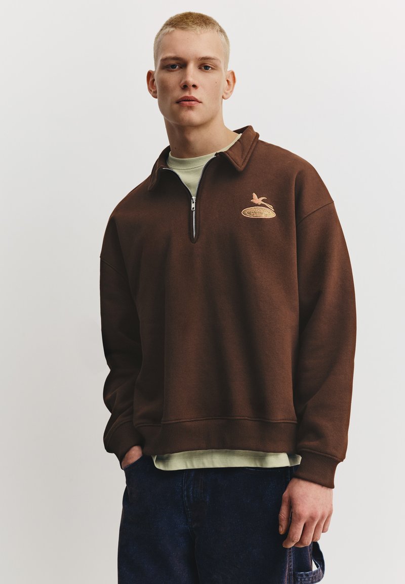 Brun sweatshirt med halv lynlås og lysegrøn undertrøje. Har en broderet logo på brystet og ribstrikkede manchetter. Blød stoftekstur.