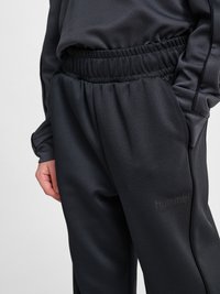 Osoba nosząca czarne elastyczne spodnie z elastycznym pasem oraz dopasowany długi rękaw z rękami w kieszeniach, prezentująca logo marki hummel na spodniach.