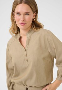 Chemisier beige avec un col volanté, une patte de boutonnage et des manches longues. Fabriqué en tissu lisse avec une texture subtile et une coupe décontractée.