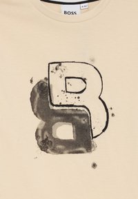 Beige T-skjorte med stor svart bokstav "B" med akvarell-effekt og flekker i grått. Myk stoff med rund hals design.