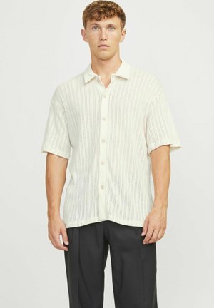 Camisa de botones de manga corta en un ligero color beige, con un diseño texturizado y acanalado que presenta recortes verticales, combinada con pantalones oscuros.
