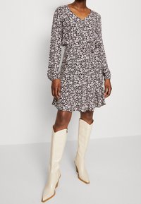 Blumenkleid mit langen Ärmeln, V-Ausschnitt, geraffter Taille und gewelltem Saum im Muster von Schwarz, Weiß und Lila. Kombiniert mit knielangen cremefarbenen Stiefeln.