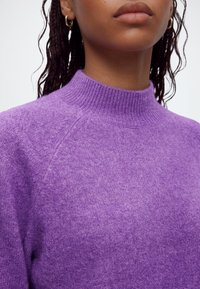Gros plan d'une personne portant un pull en maille violet à col montant et de petites boucles d'oreilles en or avec des cheveux tressés visibles.