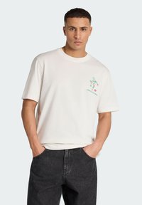 Uomo con t-shirt bianca con una piccola figura pixelata multicolore e la scritta "ITALIA 90", abbinata a jeans neri, mani nelle tasche.