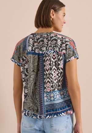 Femme aux cheveux courts et bruns portant un chemisier coloré à manches courtes et un jean bleu clair, vue de dos.