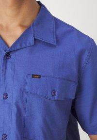 Kortärmad skjorta med krage i starkblått tyg med en ficka och lila knappar. Har en liten logotags på bröstfickan.