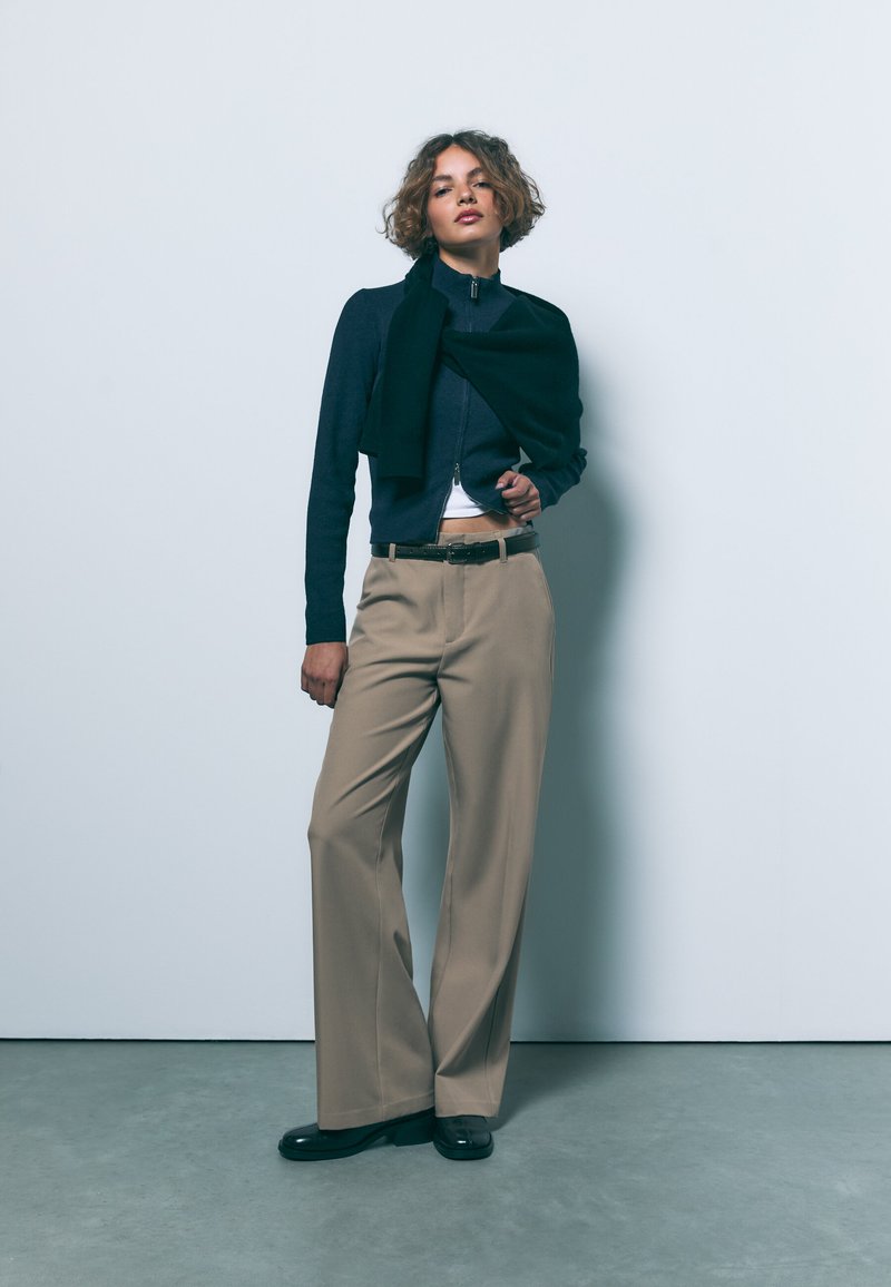 Laivaston sininen vetoketjullinen neulepaita kerroksittain mustan huivin kanssa, yhdistetty beigeihin leveälahkeisiin housuihin ja mustiin kiiltäviin kenkiin, yksinkertaista taustaa vasten.