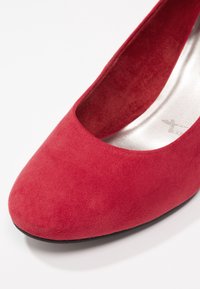 Gros plan d’une chaussure plate en daim rouge pour femme avec un bout arrondi et une semelle intérieure blanche rembourrée sur une surface blanche.