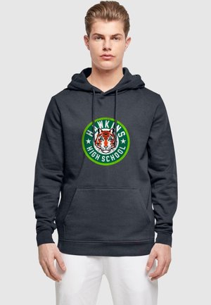 Mørkegrå hættetrøje med lomme foran. Har et cirkulært grafik af en tiger samt teksten "HAWKINS HIGH SCHOOL" i grøn og hvid.