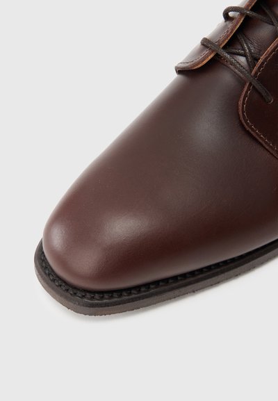 Sapato de couro castanho com biqueira arredondada, textura lisa e design com atacadores; apresenta sola costurada e salto ligeiramente elevado.