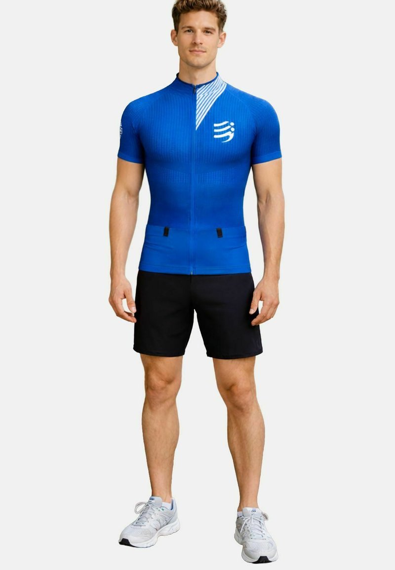Sportlicher Mann in blauem kurzärmligem Radtrikot mit Logo und schwarzer Shorts, steht mit Blick nach vorn vor weißem Hintergrund.