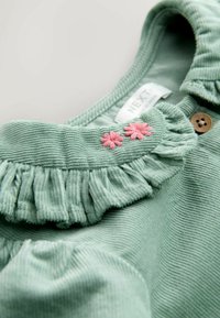 Grünes Cordtop mit rüschiertem Kragen, zwei pinken Blumenstickereien und einem Holzknopf-Akzent. Weicher Stoff und geripptes Design.