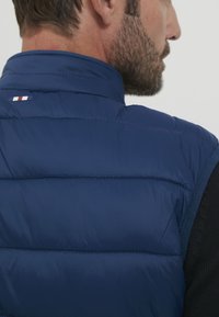 Kraljevsko modra napihnjena jakna brez rokavov z visokim ovratnikom, horizontalnim šivanjem in majhnim tridelnim poudarkom na hrbtu. Tekstura tkanine na rokavih.