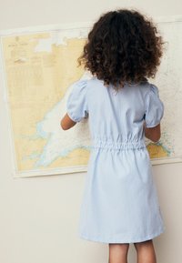 Sinivalkoinen ruudullinen mekko, jossa on kootut vyötärö, lyhyet puffihihat ja pyöristetty kaulus, päällä lapsella, joka on kasvot karttaan päin.