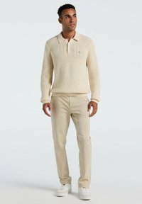 Beige gestrickter Pullover mit Polokragen, der ein strukturiertes Design aufweist. Kombiniert mit hellbeigen Hosen und weißen Sneakers.