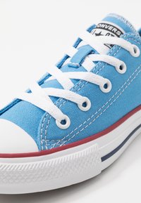 Blå canvas-sneaker med vita skosnören och gummität tåhätta. Har röd- och vitrandig sula. Förstärkta öljetter och texturerad yttersula.