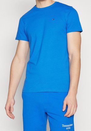 Homme portant un t-shirt bleu vif à manches courtes et un short assorti, le short est marqué du texte "Tommy Jeans ©1985" ainsi que d'un petit logo de drapeau.