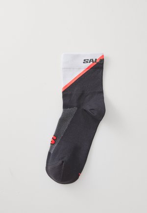 Sort og grå atletikstrømpe med hvid manchet, prydet med en rød diagonal stribe og branding. Stoffet har en glat overflade med mesh-detaljer.