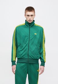 Πράσινο αθλητικό μπουφάν με κίτρινες ρίγες, ψηλό γιακά και φερμουάρ. Διαθέτει υφή με σχέδιο και ταιριαστό πράσινο παντελόνι με λογότυπο.