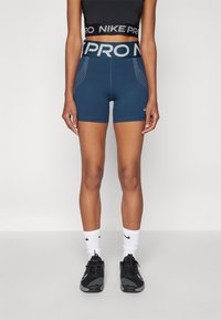 Tamsiai mėlynos Nike Pro šortai su dideliu elastingu juosmeniu, taškuotais šonų akcentais, pagaminti iš elastingos medžiagos, derinami su juodais sportiniais batais ir baltais kojiniais.