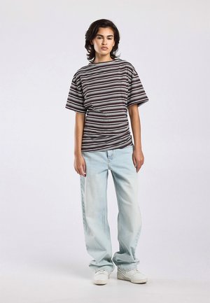 Persona joven de pie, con una camiseta holgada a rayas marrón y blanca, vaqueros de pierna ancha azul claro y zapatillas blancas, fondo neutro.