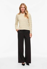 Jeune femme portant un pull beige à motifs, un pantalon large noir et des talons noirs, debout avec une main dans la poche sur un fond blanc.