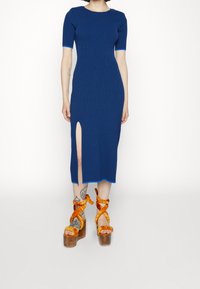 Sukienka dzianinowa midi w kolorze niebieskim, z krótkimi rękawami i bocznym rozcięciem; z kontrastującymi jasnoniebieskimi akcentami. W połączeniu z masywnymi, plecionymi sandałami na platformie.