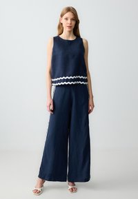 Marineblaues Zweiteiler-Outfit aus Leinen. Ärmelloses Oberteil mit weißem wellenförmigen Besatz, kombiniert mit lockeren, weiten Hosen mit seitlichen Streifen.