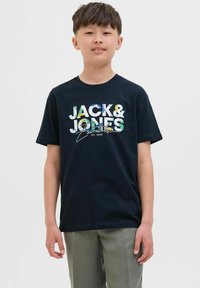 Crna majica s kratkim rukavima s šarenim grafičkim prikazom "JACK & JONES Essentials" s raznolikim teksturama; uparen s svijetlozelenim hlačama.