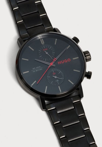 Montre bracelet en métal noir avec aiguille des secondes rouge, sous-cadrans jour et date, index des heures argentés et logo HUGO sur cadran bleu foncé.