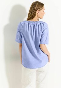 Himmelblaues, kurzärmliges Oberteil mit gerafftem Kragen, glatter Textur und abgerundetem Saum, kombiniert mit weißen Hosen. Rückansicht.