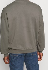 Sudadera gris de cuello redondo con mangas largas, puños y dobladillo acanalados. La parte posterior presenta una textura suave y un detalle de costura mínima. Combinada con jeans azules.
