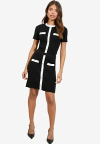 Black ribbed dress featuring short sleeves, white button accents, and front pockets. The skirt has a fitted shape and a straight hem.