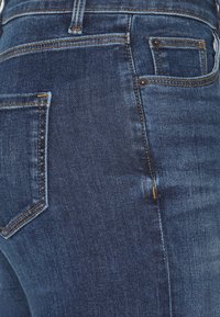 Jeans azul oscuro con una textura suave, que presentan costuras de contraste, un diseño clásico de cinco bolsillos y un ligero desgaste en la tela.