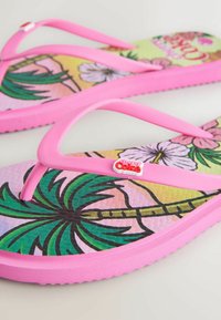 Rožinės šlepetės su tropinių gėlių ir palmės medžių raštais bei „Cherry Coke“ logotipu ant dirželių guli ant šviesios dangos.