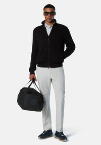 Schwarze, gerippte Zip-Jacke über einem grauen Poloshirt, weiße Hose, schwarze Sporttasche und blaue Turnschuhe. Das Model trägt dunkle Sonnenbrillen.