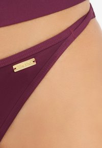 Burgunderfarbene Bikini-Hose aus glattem Stoff, mit einem Goldton-Logo-Akzent und dünnen Seitenbändern mit genähten Details.
