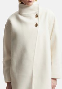 Cappotto bianco in lana con colletto alto, chiusura frontale asimmetrica e due bottoni decorativi dorati. Tessuto morbido, vestibilità oversize.
