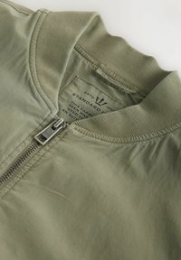 Hellolivgrüne Bomberjacke mit geripptem Kragen und silbernem Reißverschluss, teilweise geöffnet, sodass das markierte Innenetikett mit Text sichtbar ist.