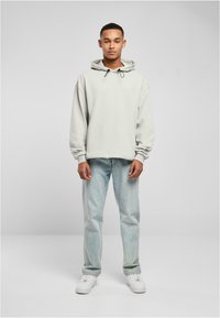 Sudadera gris claro con capucha ajustable, combinada con jeans de denim azul claro y zapatillas blancas, de pie sobre un fondo neutro.