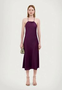 Tânără îmbrăcată cu o rochie midi violet cu bretele, ținând o poșetă mică de culoare verde deschis, stând în fața unui fundal simplu deschis.